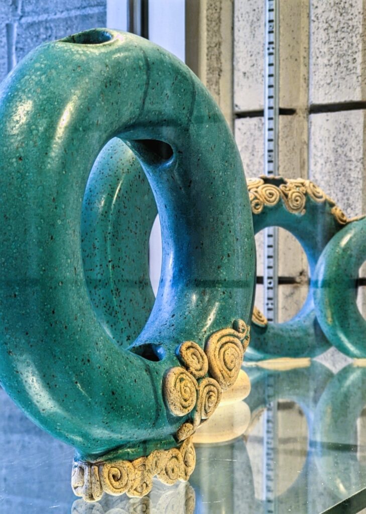 A shot along a glass shelf holding three matte turquoise "donut" vases with raw clay embellishments.