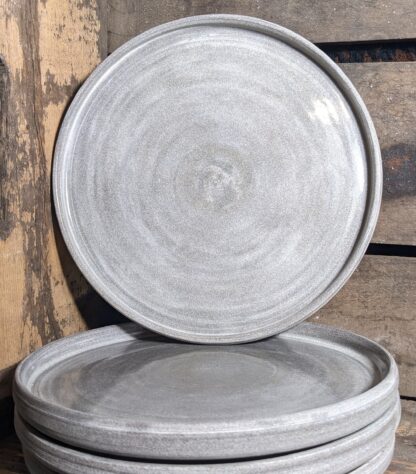 stack of 4 plates, with one plate standing up to show the food surface of the plate. Plates are made of PSH dark granite clay body and are a variable gray colour, glazed in a clear glaze.